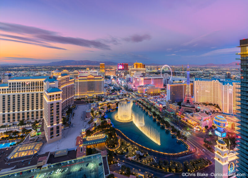 Sunset view of the Las Vegas Strip from a luxury hotel room at the Cosmopolitan. The image captures the Bellagio Fountains, iconic hotels like Paris, Caesars, Harrah's, Flamingo, and the Venetian, with rare red clouds over the skyline.