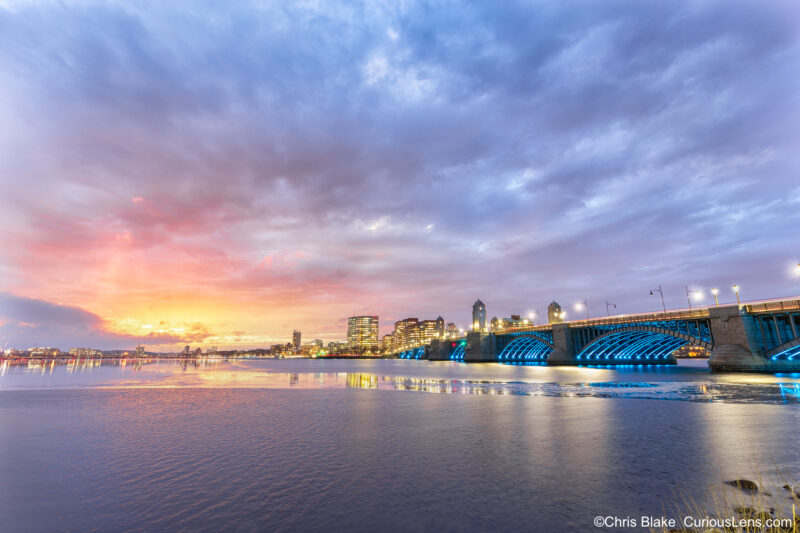 Longfellow Bridge at sunset with Charles River half-covered in ice, blue lights under the bridge, and fiery sky over Cambridge.