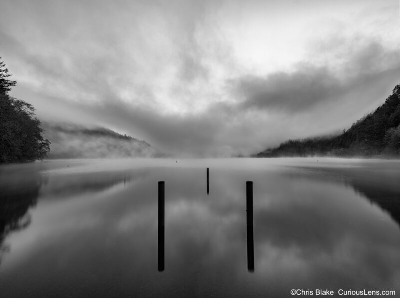 Lake Crescent at sunrise with deep fog, mist rising from the water, three dock pillars centered in the frame, and dark mountains on each side.