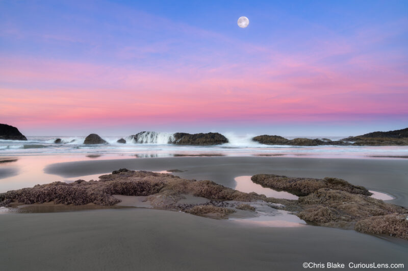 Sunrise at Seal Rock State Park with the moon setting over the Pacific Ocean, tide crashing over rocks, and warm red glow from reflected light.