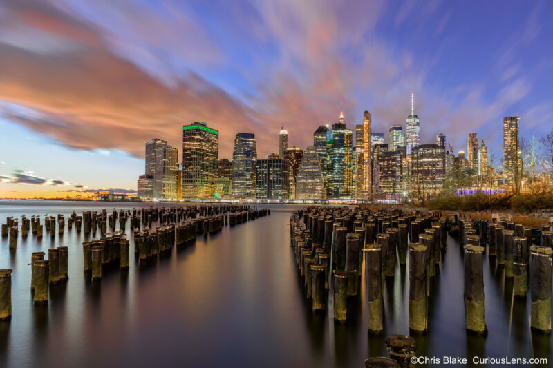 Sunset over lower Manhattan from Brooklyn Bridge Park with remains of old Pier 1, iconic skyline, One World Trade Center, and vibrant sky.