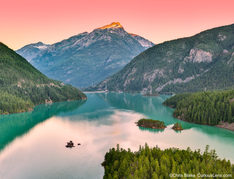 Diablo Lake in North Cascades National Park with turquoise water, dam, power lines, and snowcapped mountains at sunrise.