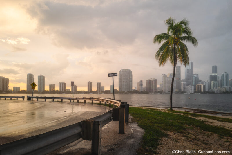 Miami skyline after summer thunderstorm with winding road, glistening guardrail, palm tree, and clearing sky revealing vibrant red sunset.