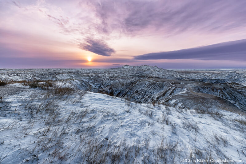 Sunrise at Panorama Point in Badlands National Park with snow, rock, and grass layers, colorful clouds, and warm sunlight breaking through storm clouds.