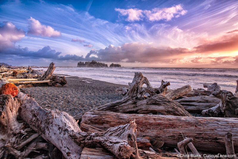 Rialto Beach in Olympic National Park with vibrant sky, colorful clouds, massive logs on the rocky beach, and a lone island in the distance.