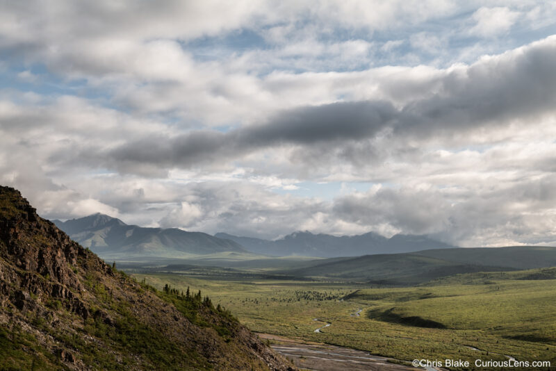 Denali National Park with a looming mountain, valley of meandering rivers and lush hills, distant mountain range, and sky with remarkable clouds and scattered light.