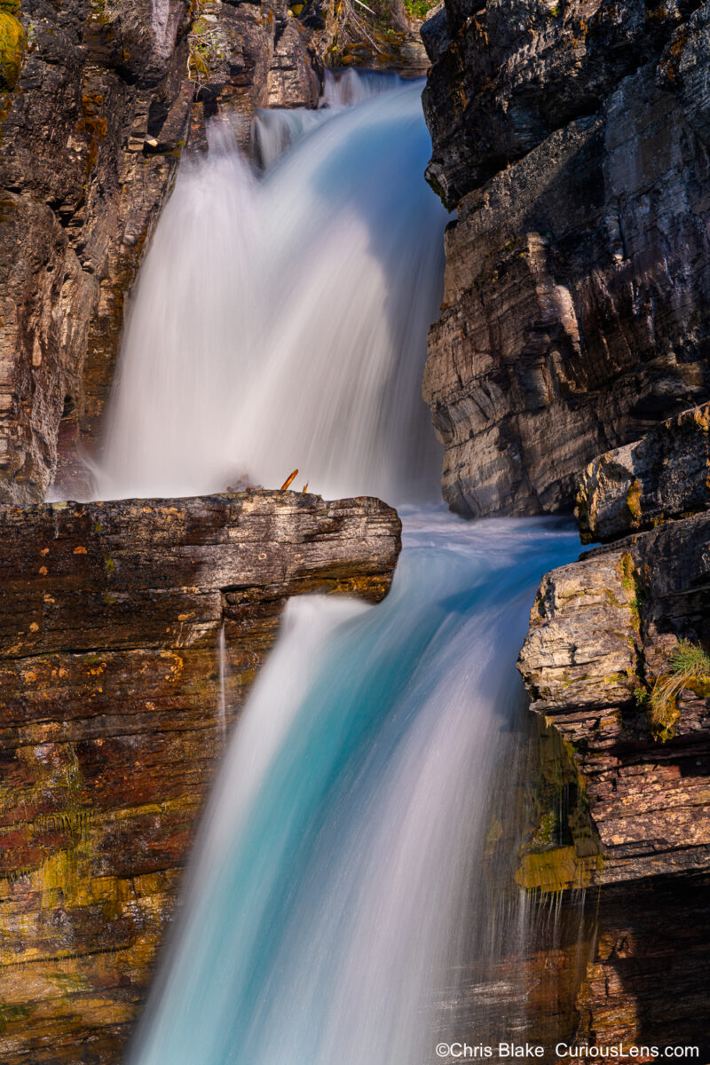 Saint Mary’s Falls in early morning light with strong flow, blue and teal water, contrasting rocks, and green glowing moss."