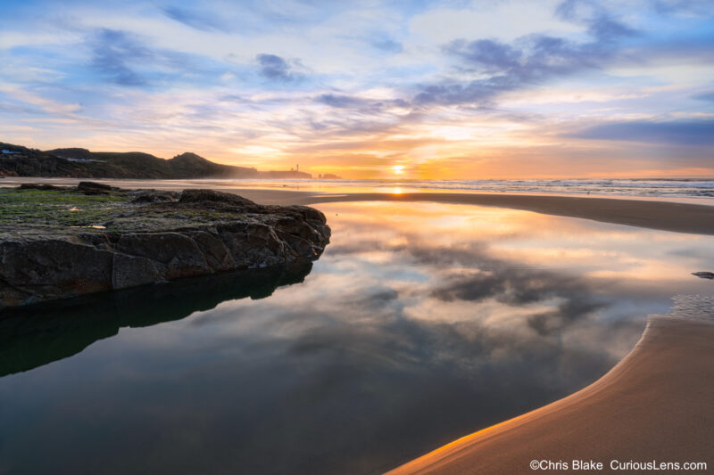 Sunset on the Oregon coast with glowing sand, sky reflecting in water, and yellow sky behind a distant lighthouse.