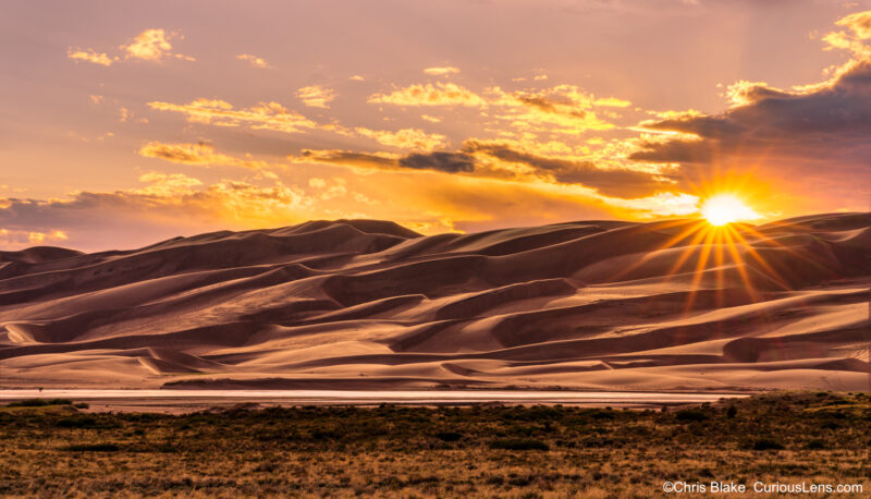 Sunset at Great Sand Dunes with sun star, warm glow, deep shadows, Medano Creek, and tiny hikers for scale.