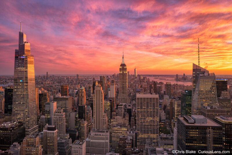 Sunset view from Rockefeller Center Observatory with Empire State Building, One World Trade Center, glowing Hudson River, and city skyline lit up.