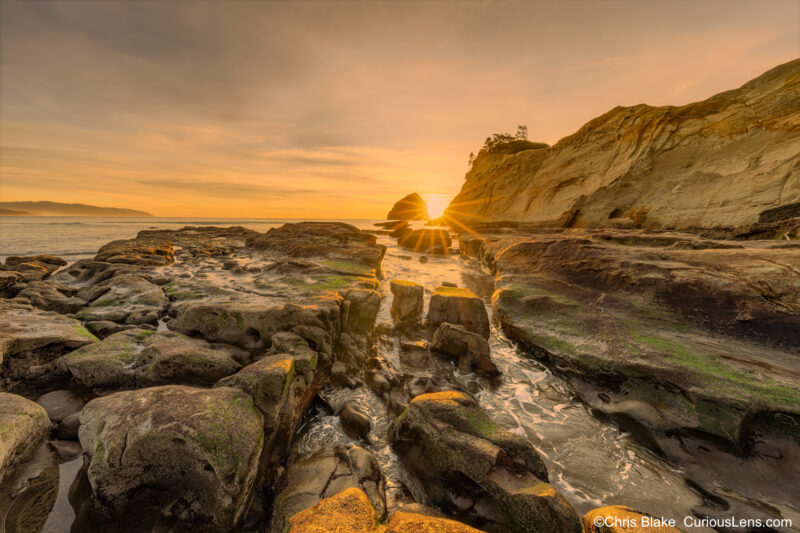 Sunset at Cape Kiwanda with tide pools, sandstone cliffs, dunes, Chief Kiawanda Rock, and sun star with warm light on the rocks.