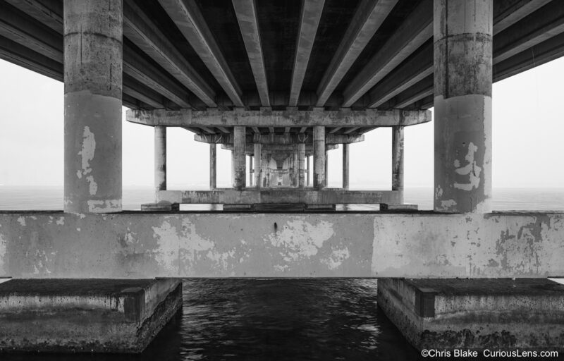 Powell Bridge during summer tropical storm with rain pouring, lightning, bridge's symmetry against weathered concrete, and calm ocean.