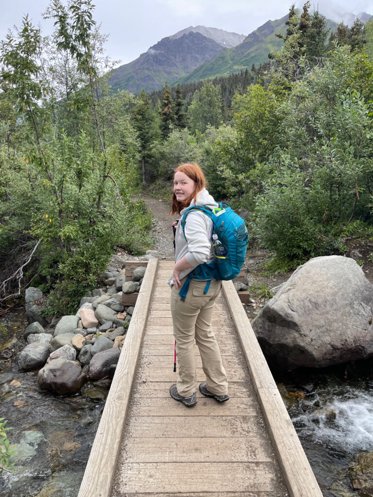 Cameron wearing hiking pants, long sleeve shirt and backpack, crossing a wooden bridge over the Bonnaza Creek on the Root Glacier Trail in Wrangle St. Elias National Park.