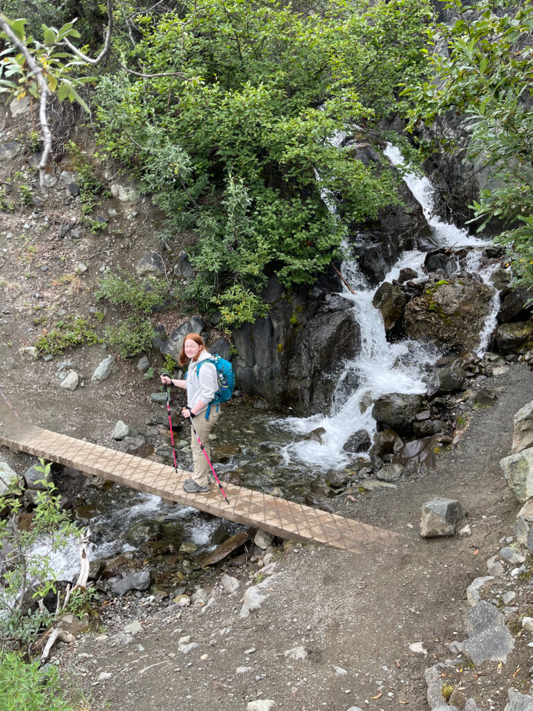 Cameron in hiking clothing and carrying a backpack on the Root Glacier Trail, walking over a very narrow board that crosses the Jumbo Creek River with waterfalls right upstream. She is looking back at me at the top of the hill and smiling. 