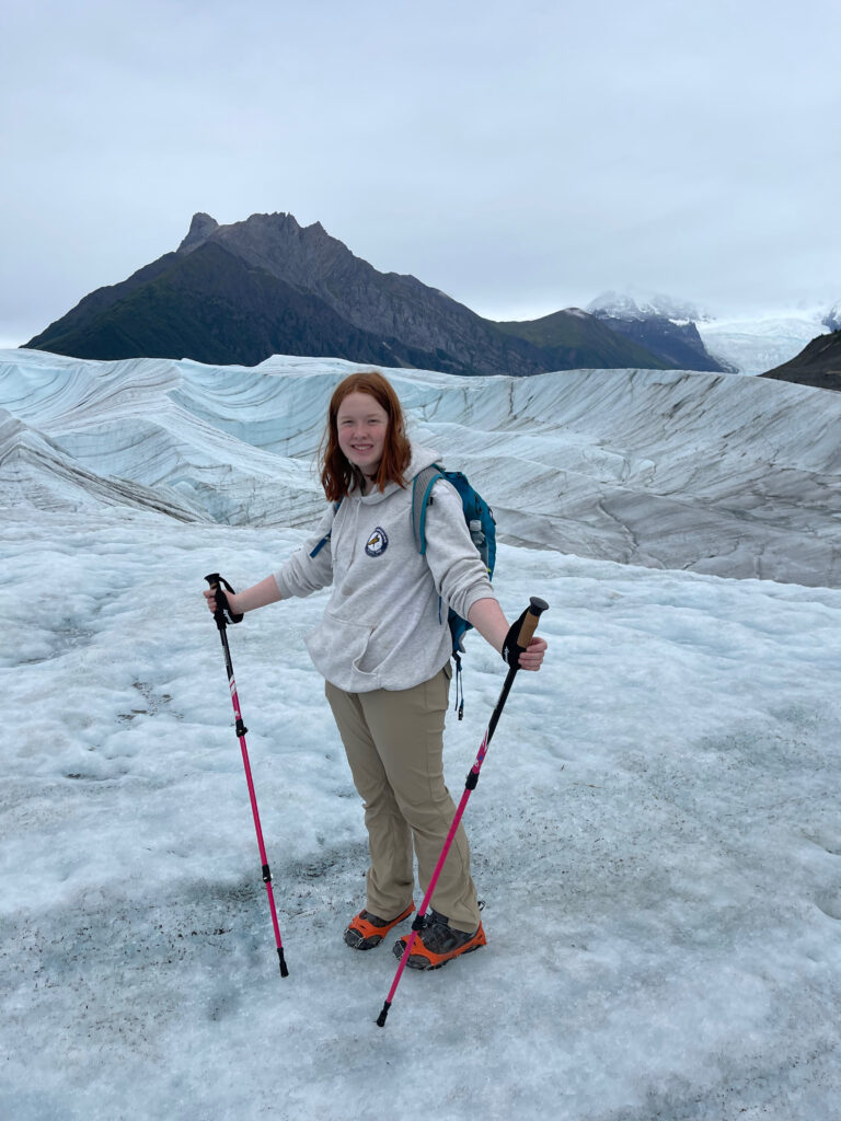 Cameron standing in the middle of the Root Glacier in Wrangle St. Elias National Park. Wearing hiking clothing, crampons on her shoes, and using trekking polls. 