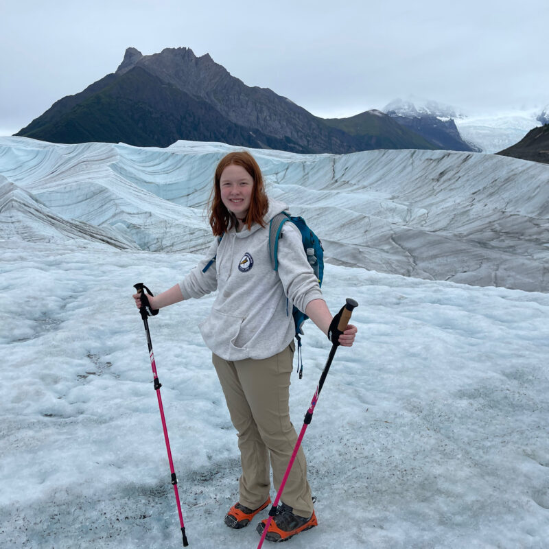 Cameron standing in the middle of the Root Glacier in Wrangle St. Elias National Park. Wearing hiking clothing, crampons on her shoes, and using trekking polls.