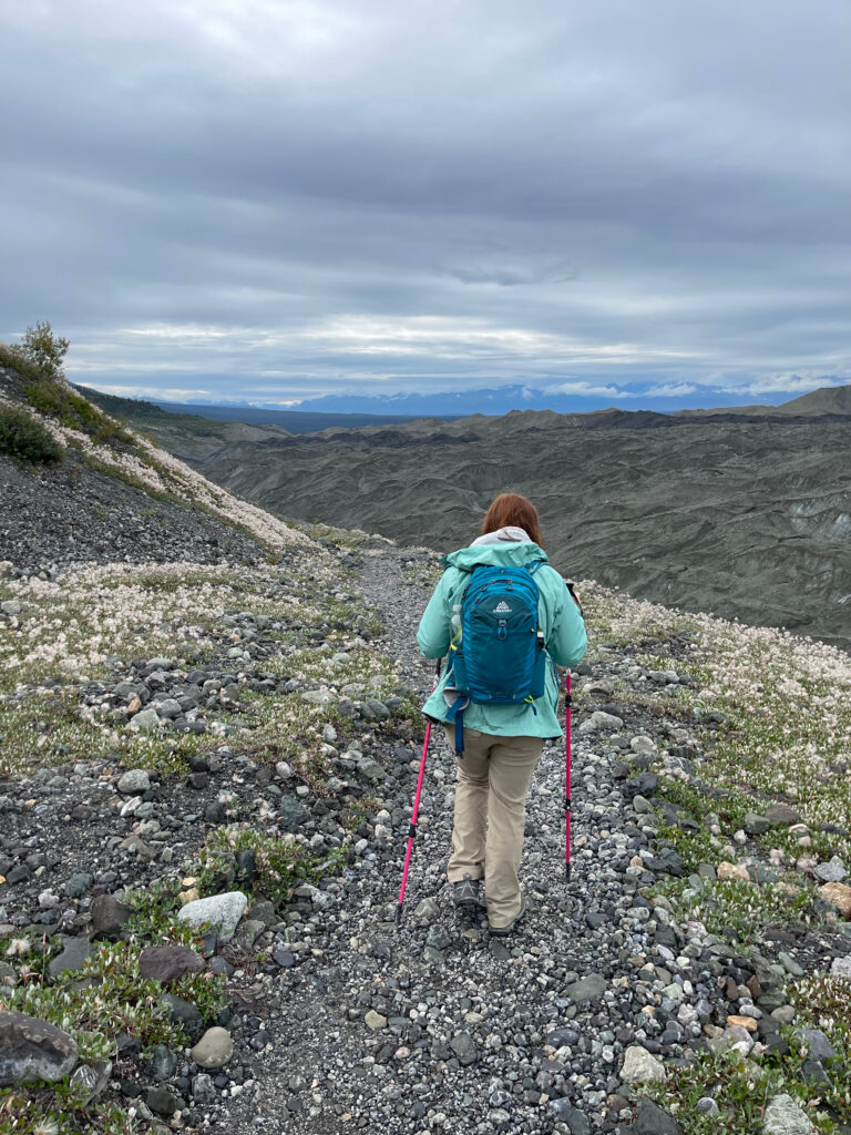 Cameron on the Root Glacier Trail in Wrangle St. Elias National Park, wearing her raincoat and carrying a backpack. She is using hiking poles as she makes her way up the steeps rocks coming back from the glacier. 