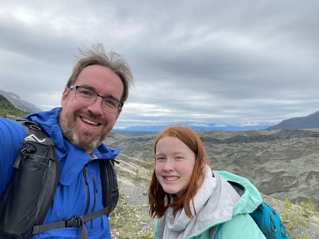 Cameron and myself pose for a photo at the top of a massive hill after hiking on the Root Glacier in Wrangle St. Elias. We both have on rain coasts and backpacks, storm clouds are overhead. 