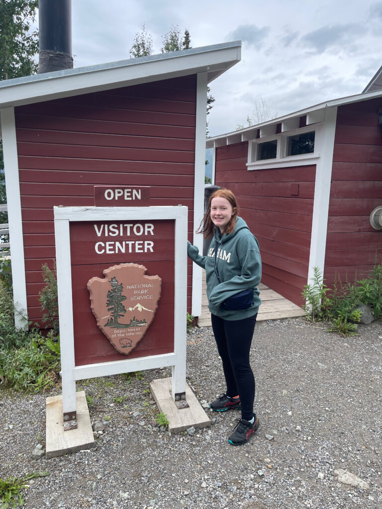 Cameron wearing a green hoodie standing next to the visitors center sign, in Kennecott, inside of Wrangell-st. Elias National Park.