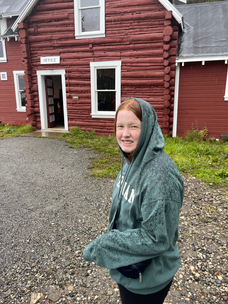 Cameron wearing a green hoodie and walking in the rain, is in front of the old Railroad Depot Station in Kennicott.