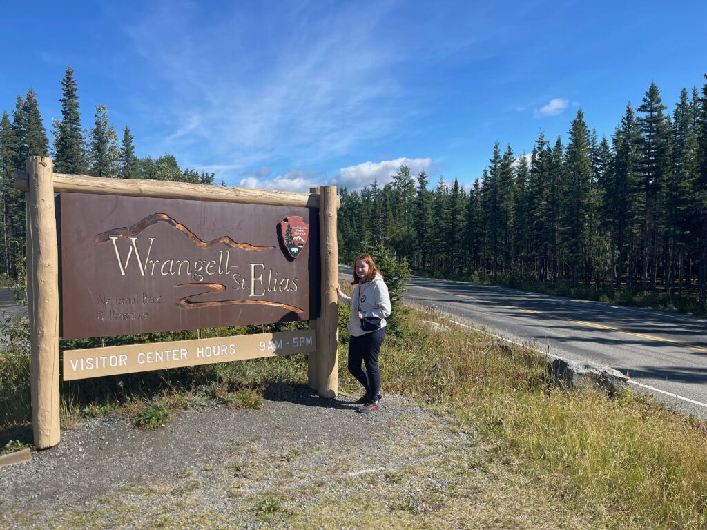 Cameron on a clear blue sky day in August standing in front of the Wrangell-St. Elias National Park sign, wearing black pants and a hoodie. 