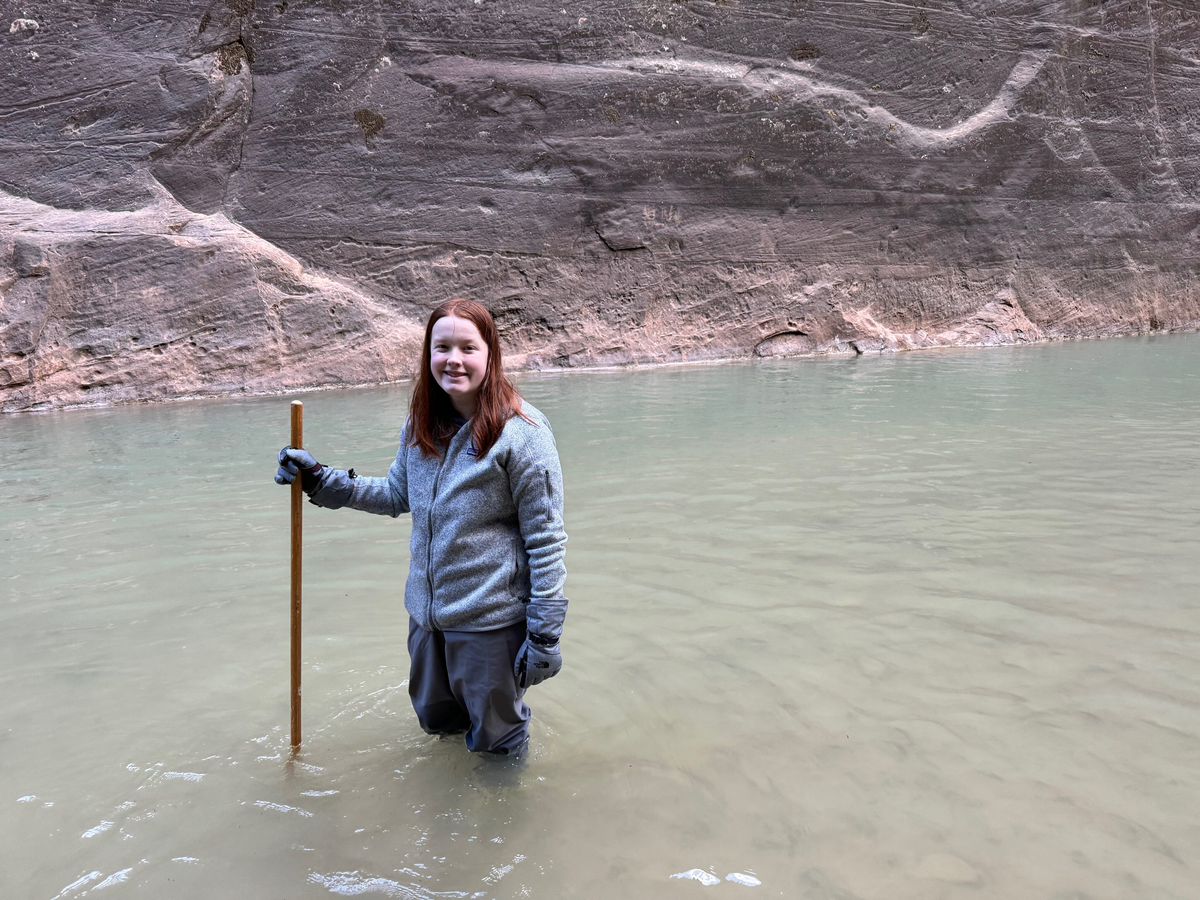 Cameron standing in the narrows, in the middle of the river, wearing a wet suit - dry bib, and a walking stick. 