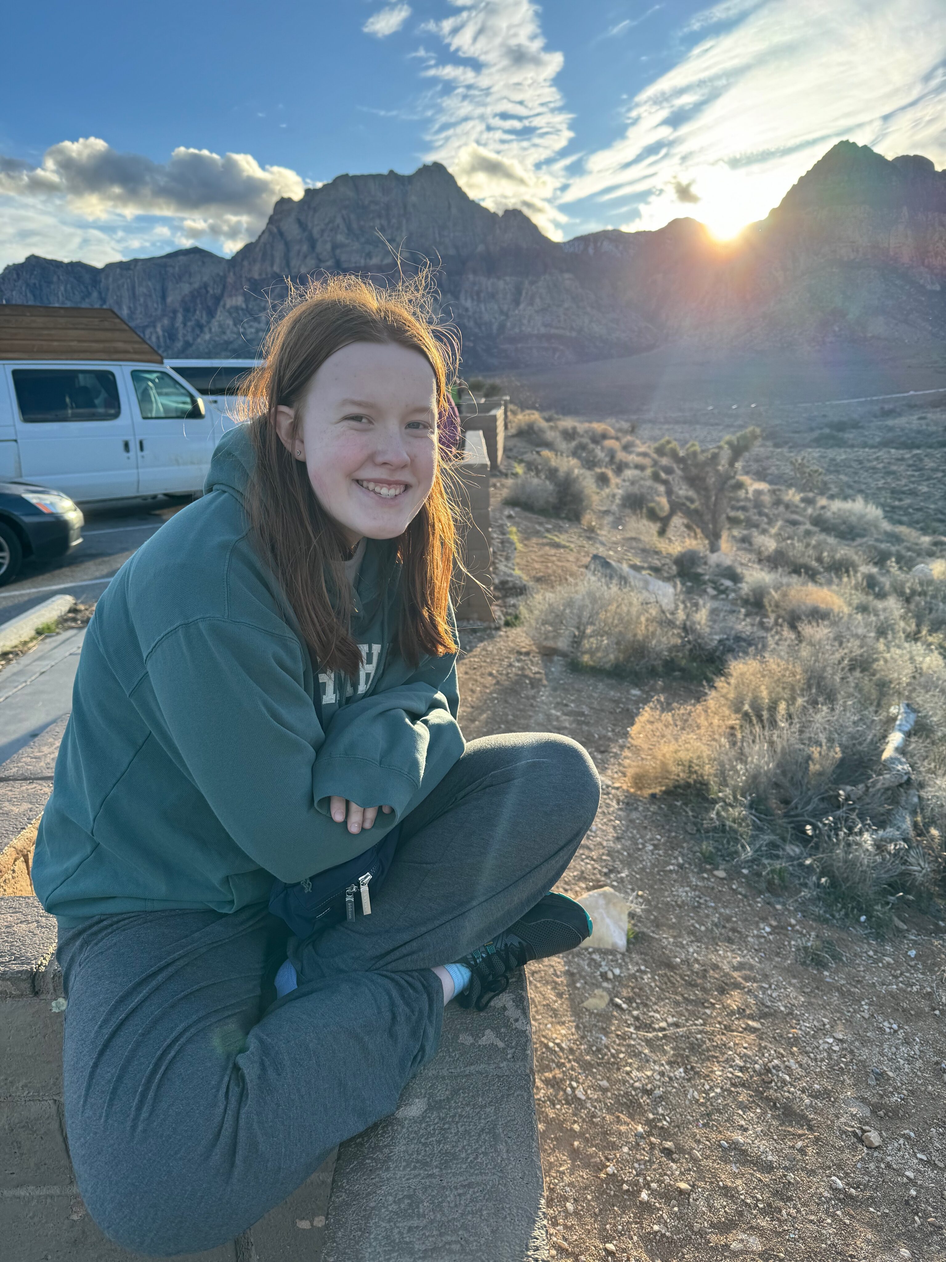 Cameron sitting cross-legged on  a wall at the Red Rocks Overlook trailhead as the last light makes it way over the mountains in the distance.