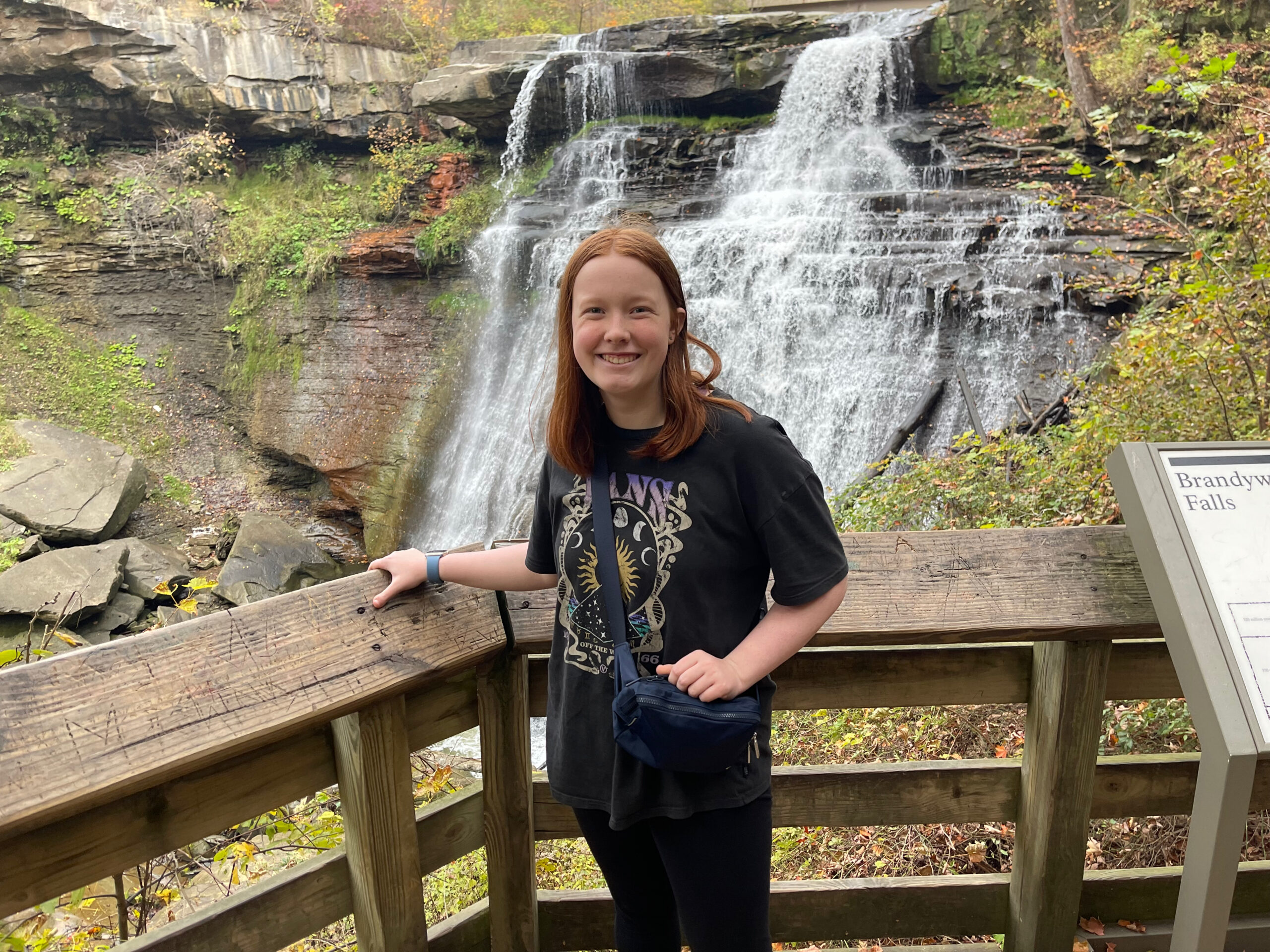 Cameron standing by the wooden railing at the base of Brandywine falls. Water rushing down the rocks and the road is above us.