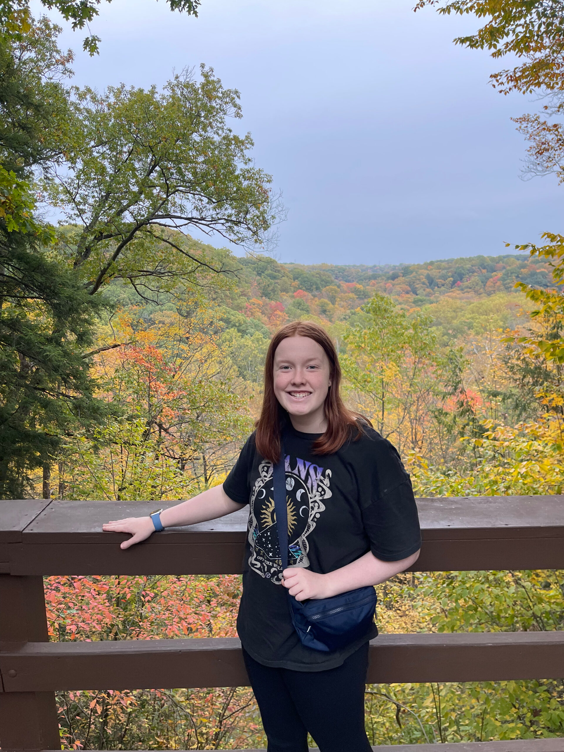 Cameron smiles as she poses at an overlook off the Gorge Parkway. A large valley full of red and yellow fall colors is behind her.