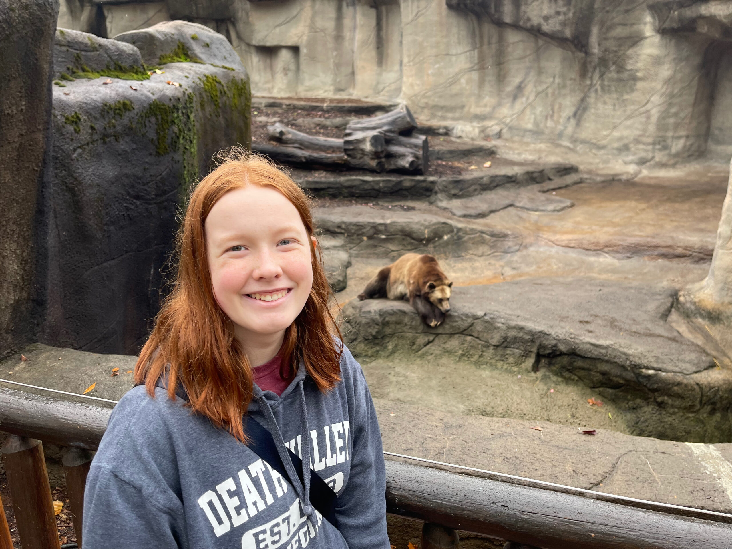 Cameron wearing a sweatshirt smiles by the railing at the MetroParks Zoo with the brown bears just behind her.