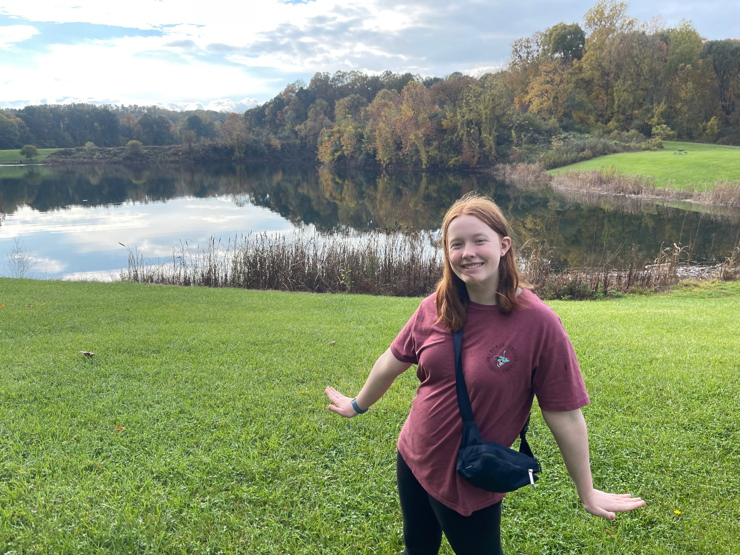 Cameron smiling with her arms out in the middle of a large grass field near Indigo Lake. The falls colors of the trees around the lake are being reflected in the water.