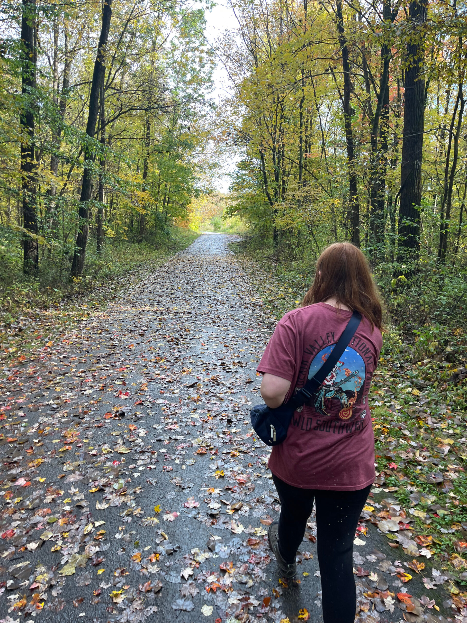 Cameron hiking down the Howe Hale Connector Trail, the ground is wet from recent rain and the trail is mostly covered in leafs. Cami is wearing a t-shirt and holding her purse.