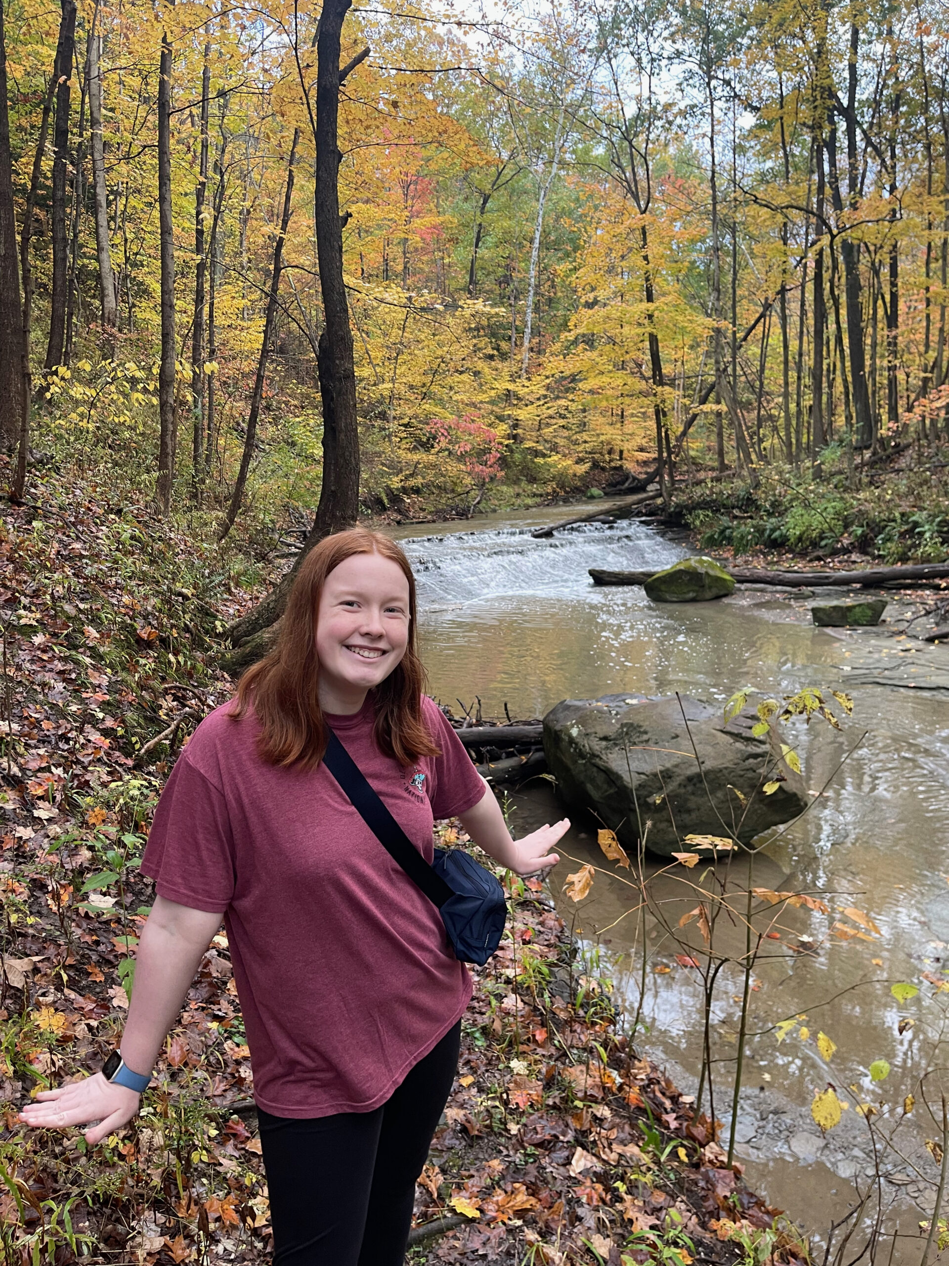 Cameron smiling wearing a death valley hoodie in the middle of the woods at the height of fall colors. There is a small but rushing waterfall and river behind her.