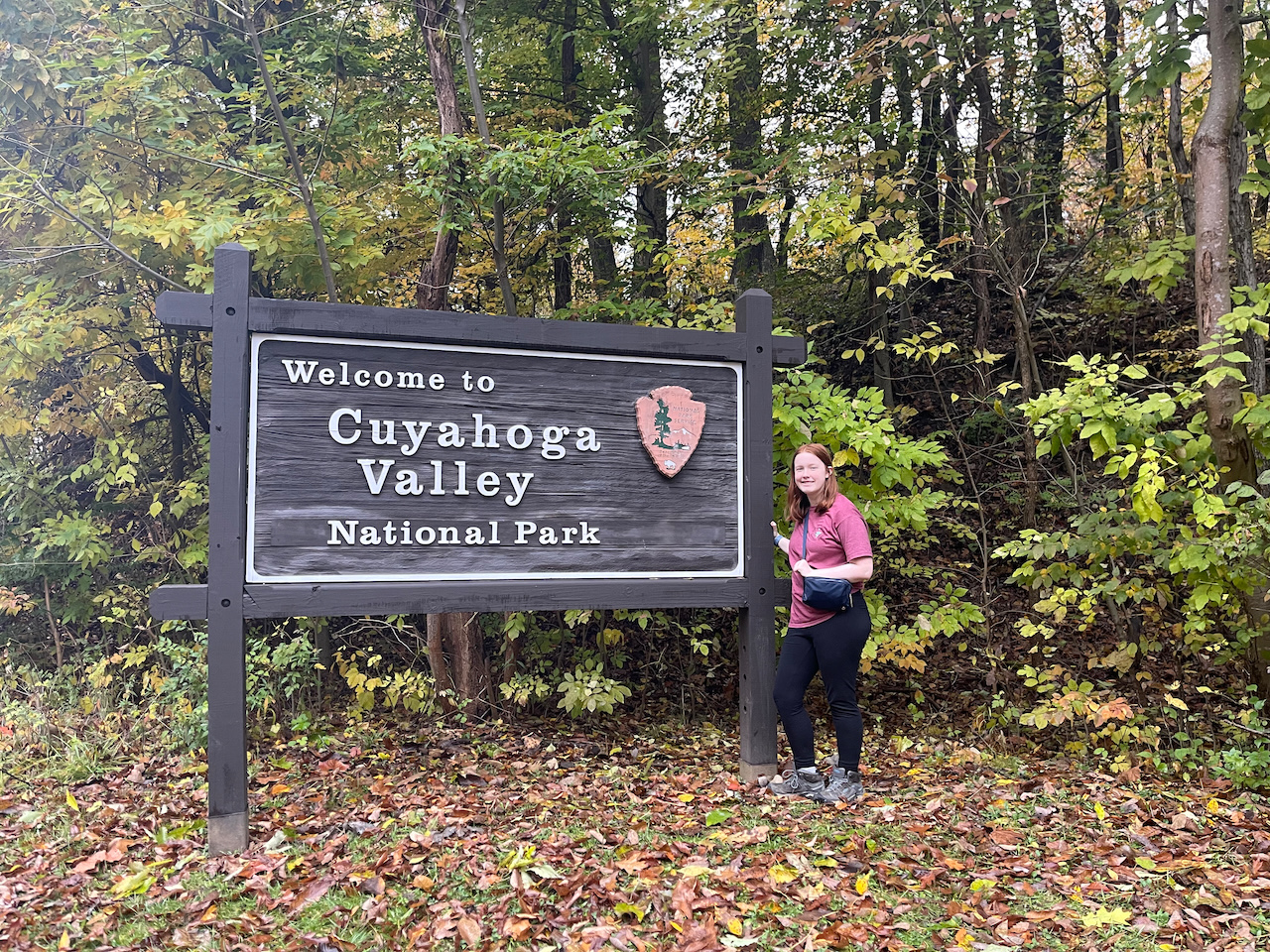 Cameron standing near the woods next to the park sign for Cuyahoga Valley National Park at the peak of fall colors.