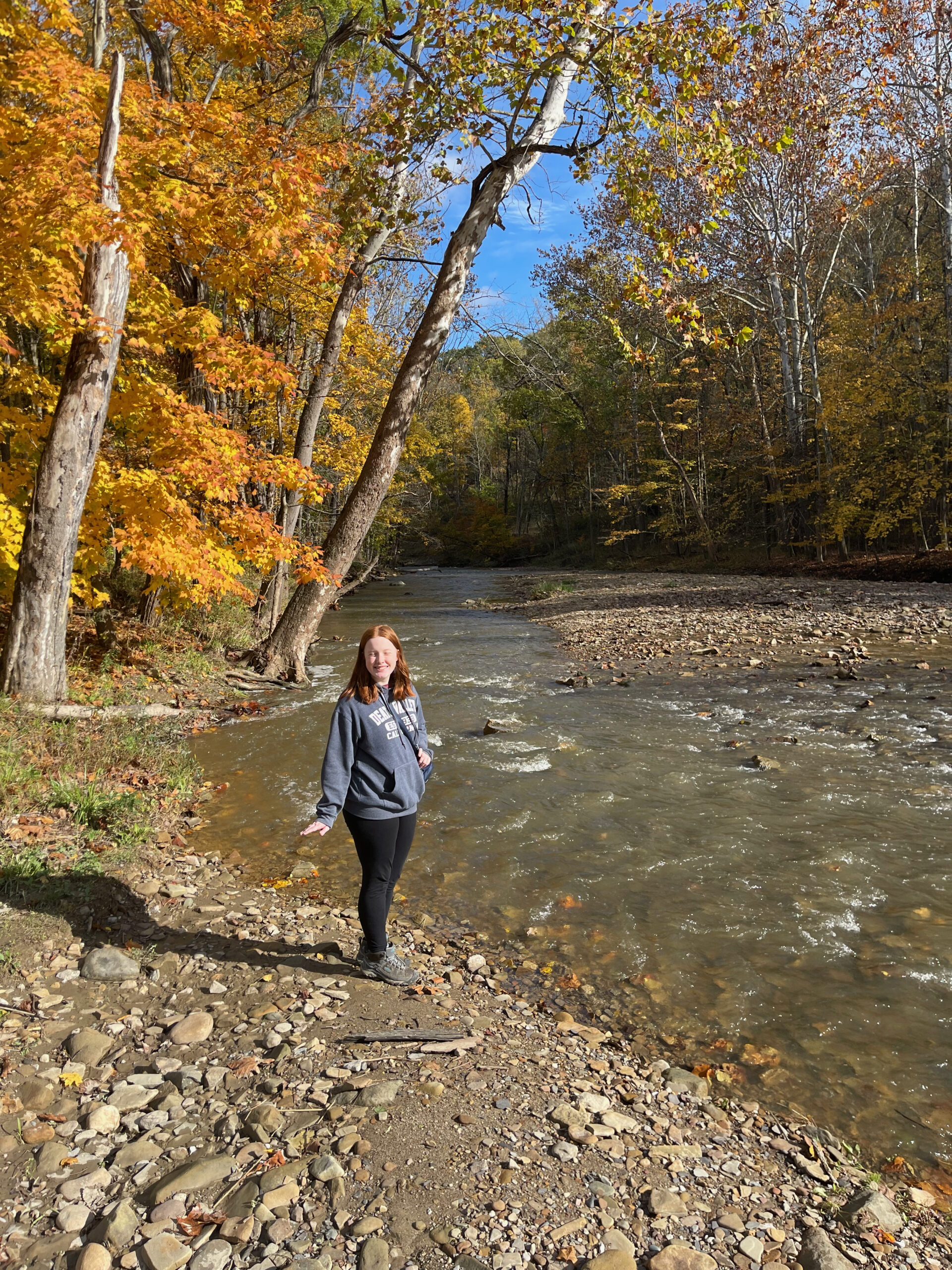 Cameron standing on the banks of the Chippewa Creek, with yellow and orange trees full of fall colors in the background.