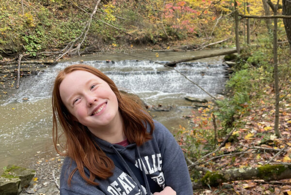 Cameron smiling wearing a death valley hoodie in the middle of the woods at the height of fall colors. There is a small but rushing waterfall and river behind her.