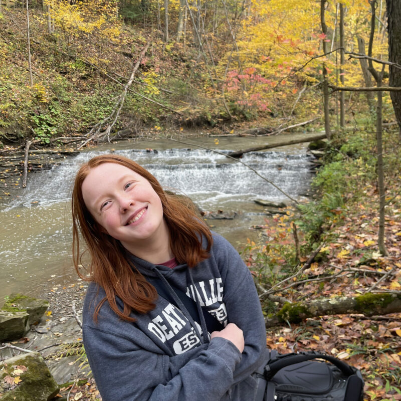 Cameron smiling wearing a death valley hoodie in the middle of the woods at the height of fall colors. There is a small but rushing waterfall and river behind her.