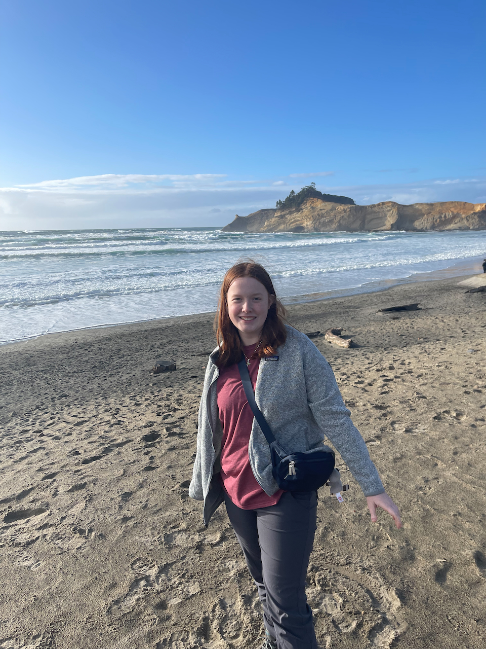 Cameron wearing hiking pants and Patagonia sweater walking on Pacific City Beach,  in the background you can see Cape Kiwanda Sand Dunes.