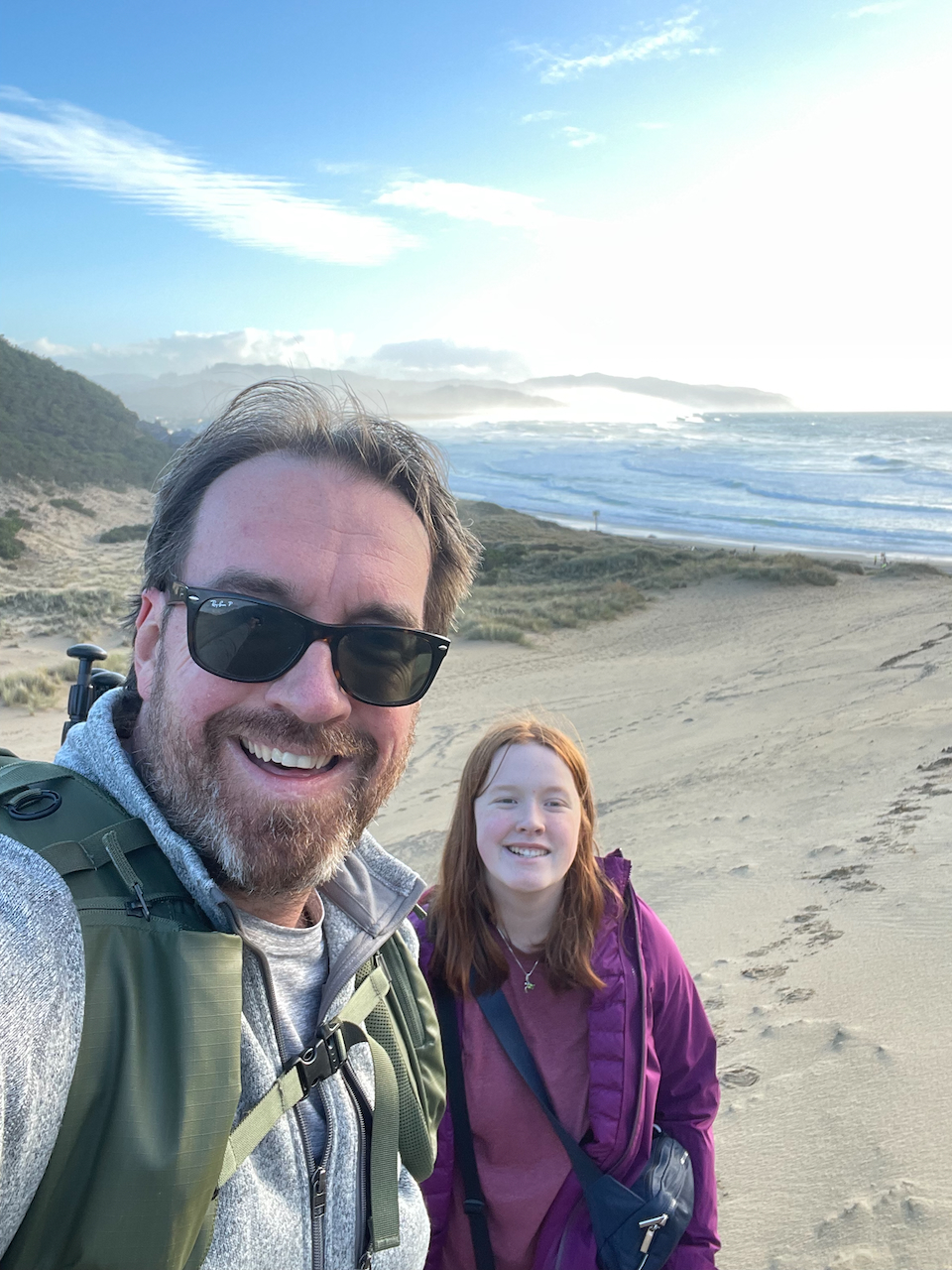 Cameron wearing a purple sweatshirt and myself wearing sunglasses and carrying a my camera backpack standing ontop of the Cape Kiwanda Sand Dunes with clouds and the ocean below us.