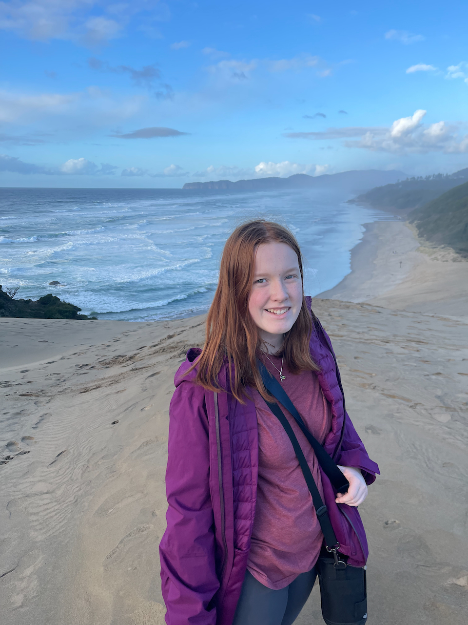 Cameron wearing a purple jacket and shirt and standing on top of the Cape Kiwanda Sand Dunes. The Pacific ocean is below us with waves crashing down.