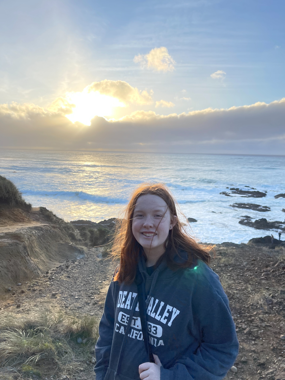 Cameron standing on rocks along the Oregon coast with a big smile on her face and the sun that's about to go behind the clouds that are on the horizon line. 