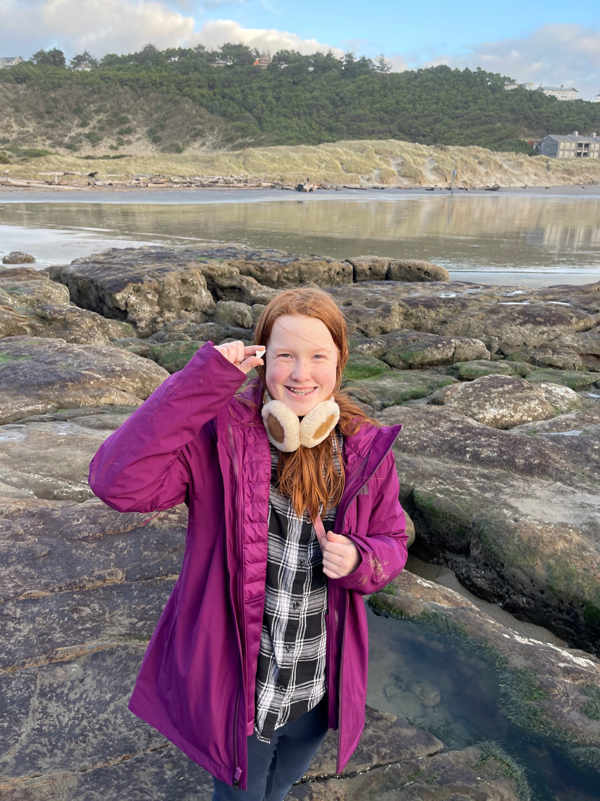 Cameron wearing a purple jacket, unzipped with earmuffs around her neck, holding up the sea glass she had just found. Standing on the rocks with a smile in Cape Kiwanda State Natural Area. 