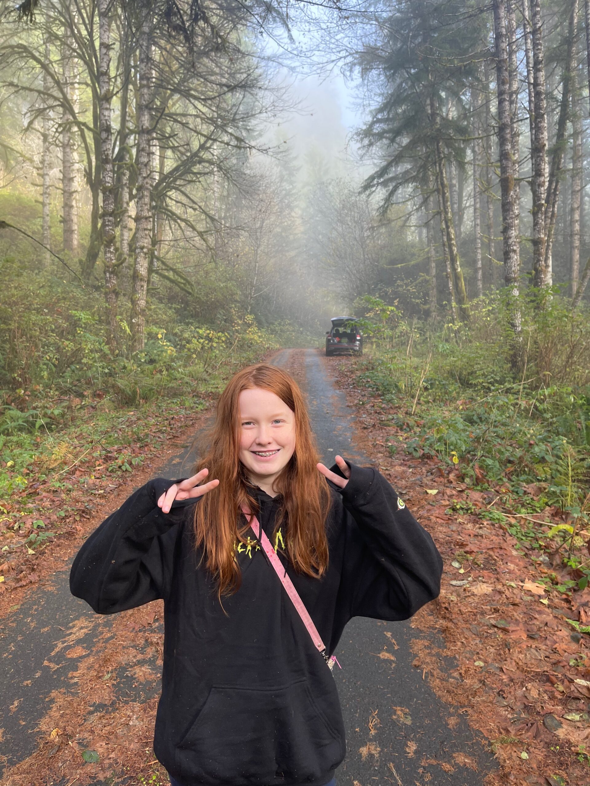 Cameron wearing a sweatshirt, with a smile on her face, making a double peace sign. Standing on a tiny road in the back country of Oregon. The fog is just breaking and you can start to see the forest.