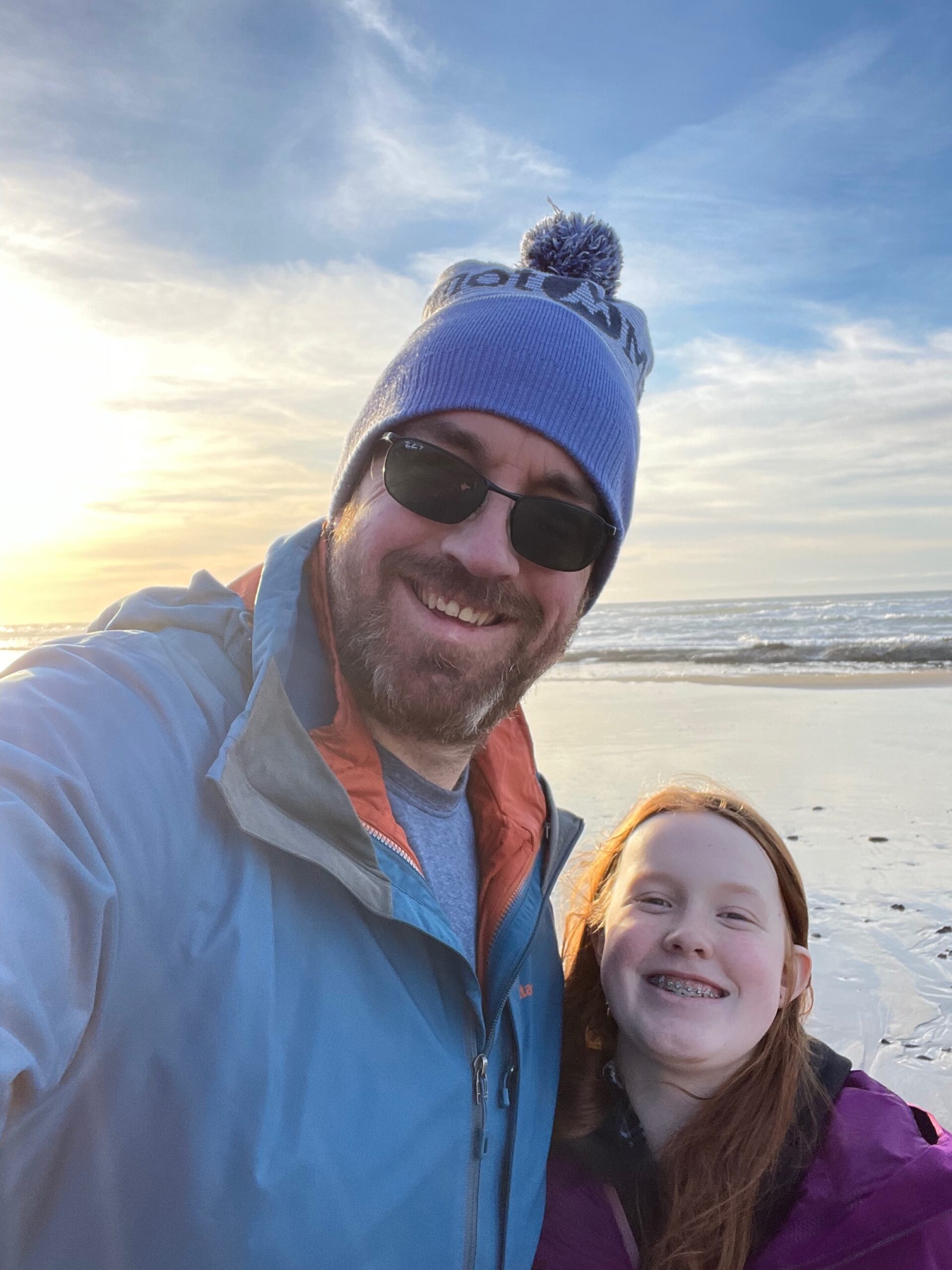 Cameron and myself, both wearing winter jackets, and me in a hat and sunglasses. Standing on Oregon Coast Beach, with a sky full of clouds and the sun just breaking through enough to light up half of Camerons face.