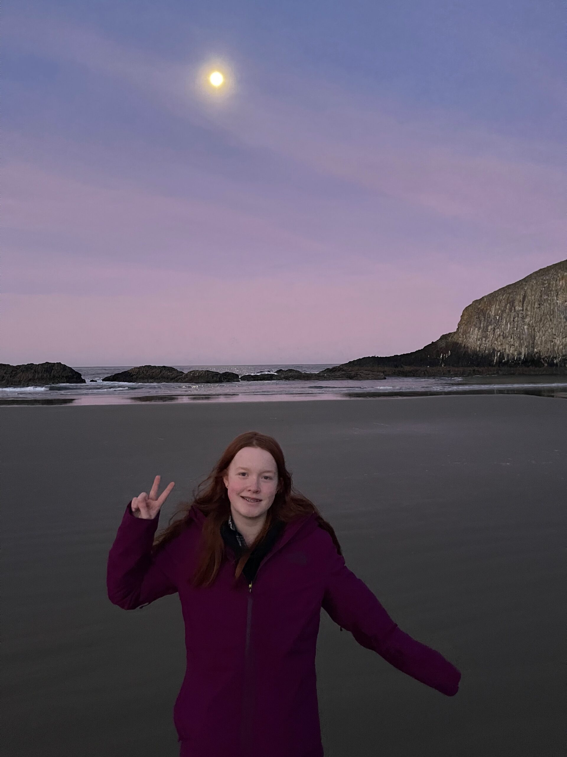 Cameron smiling making a peace sign standing on Seal Rock Beach - the sand is perfectly smooth and the moon is rising above the ocean.