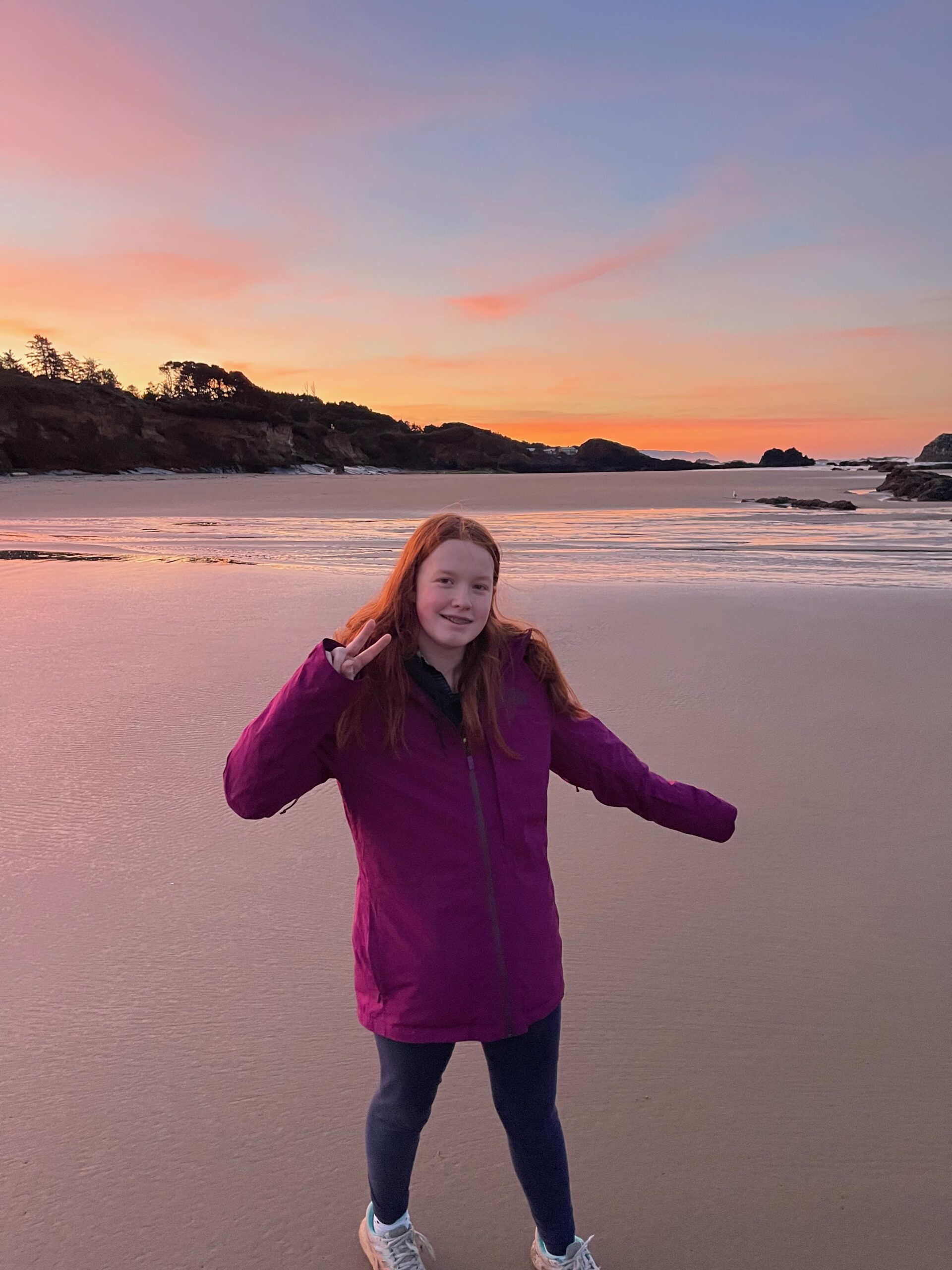 Cameron standing on the smooth sand, at sunrise at Seal Rock in Oregon. The sky is red and orange from the sunrise.