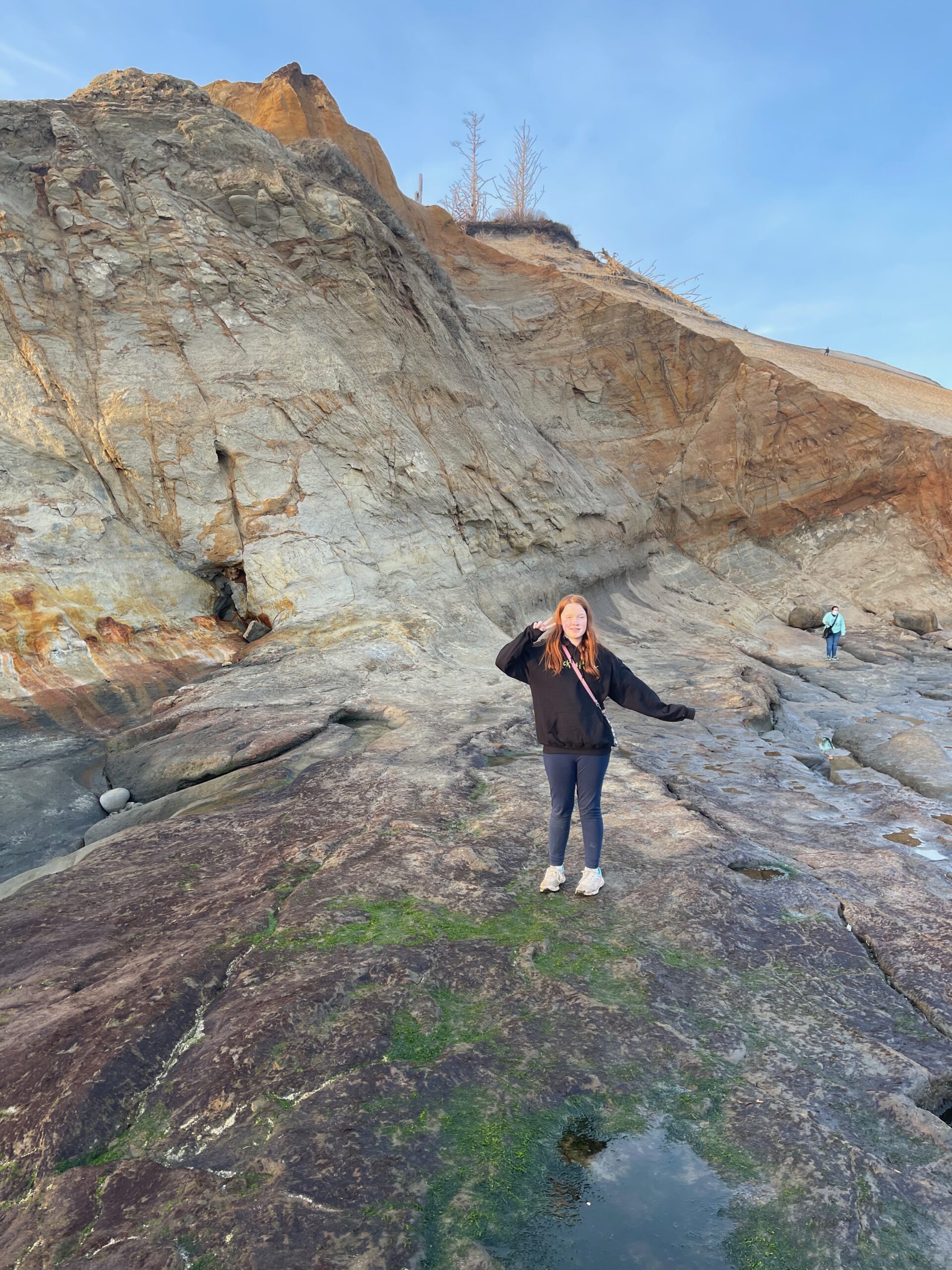 Cameron making a peace sign standing on the rocks next to the massive dunes and cliffs at Cape Kiwanda. The last light of the day is lighting her face.