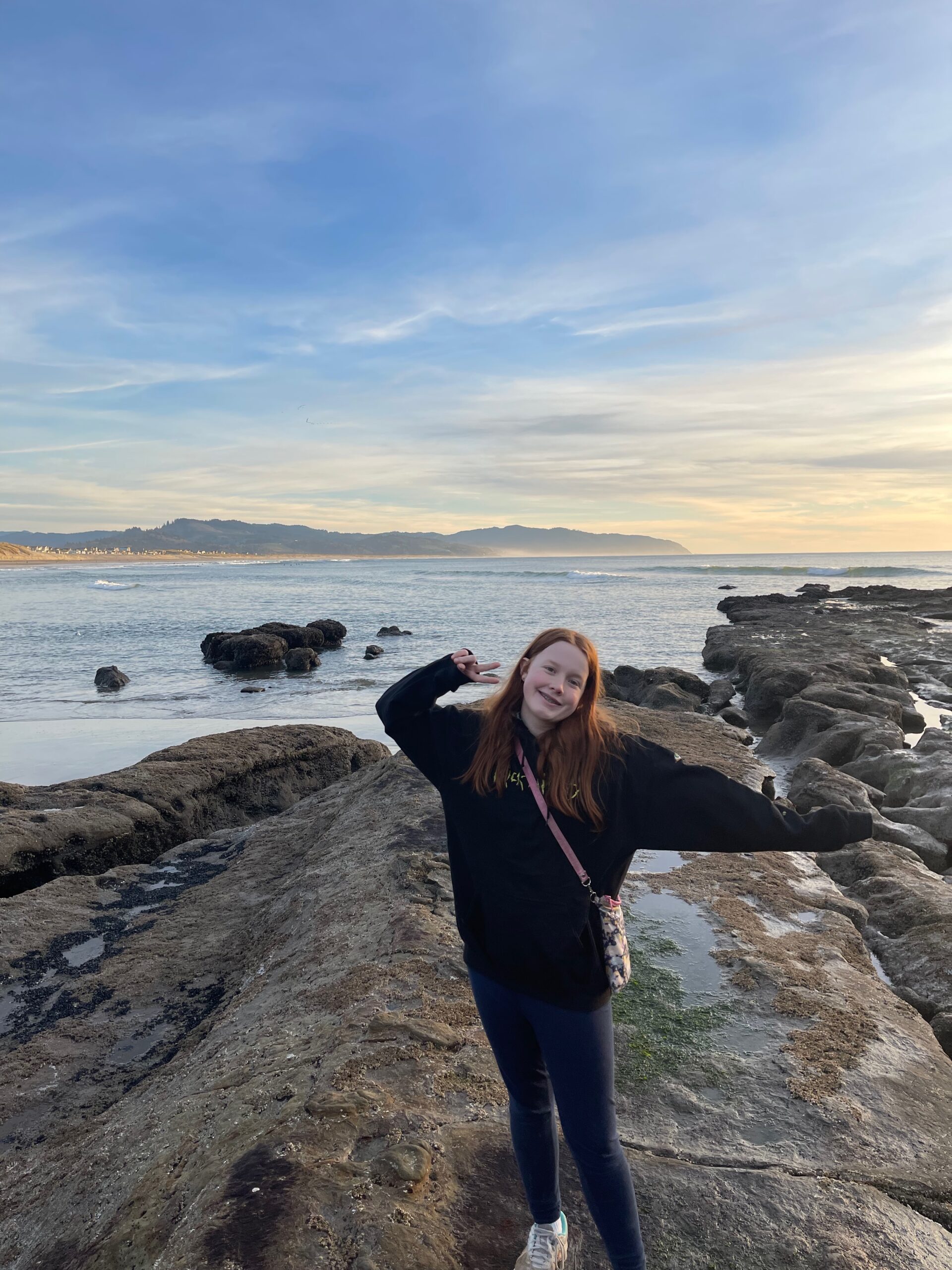 Cameron wearing a hoodie and making a peace sign, smiles as she stands on the rocks and tidal pools at low tide in Cape Kiwanda as the wave crash in the distance behind her. 