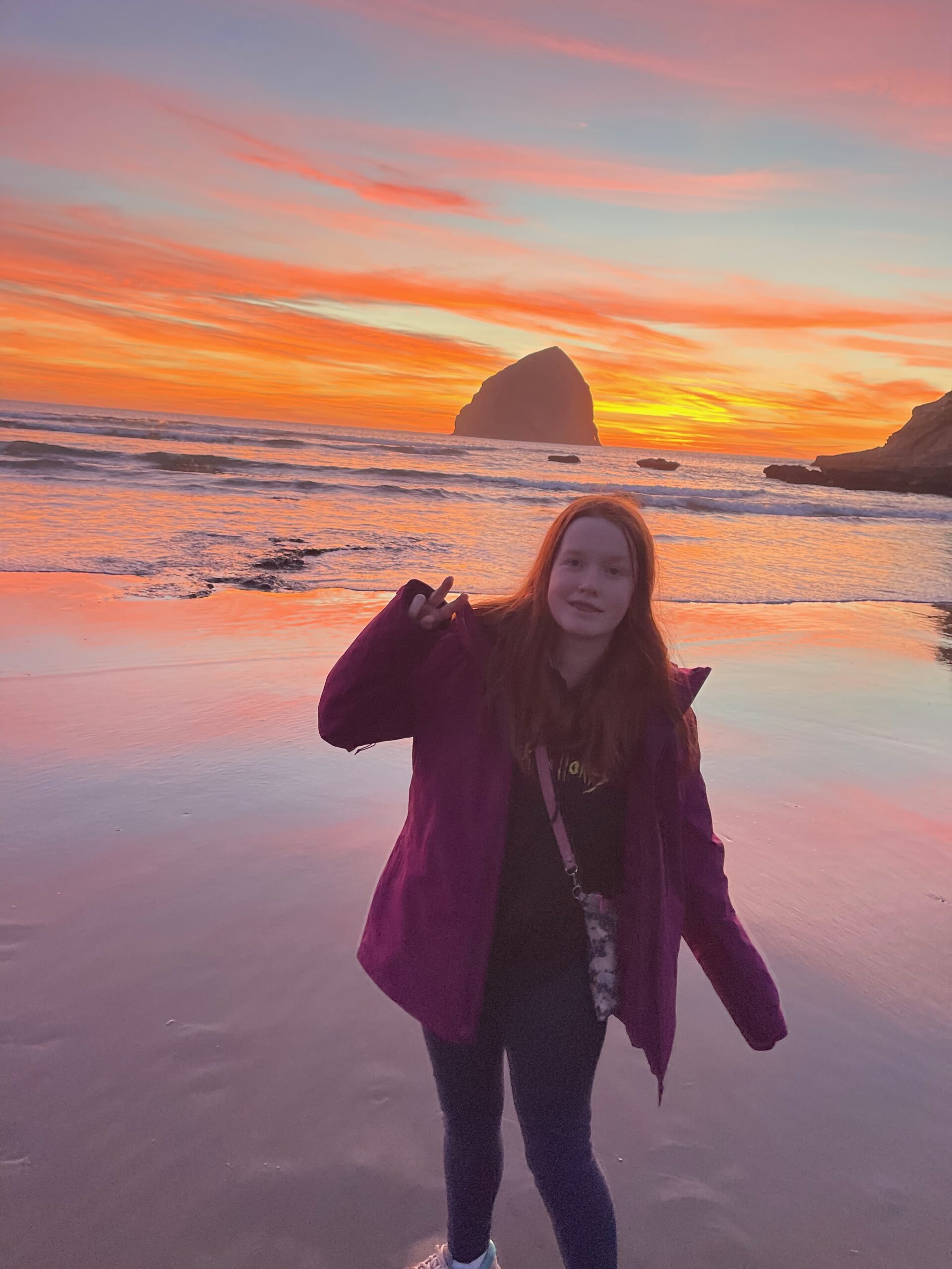 Cameron smiles and makes a peace sign at sunset at Cape Kiwanda. The sunset has light up the sky red and it's reflected in the ocean behind her. Chief Kiwanda Rock is directly behind her. 