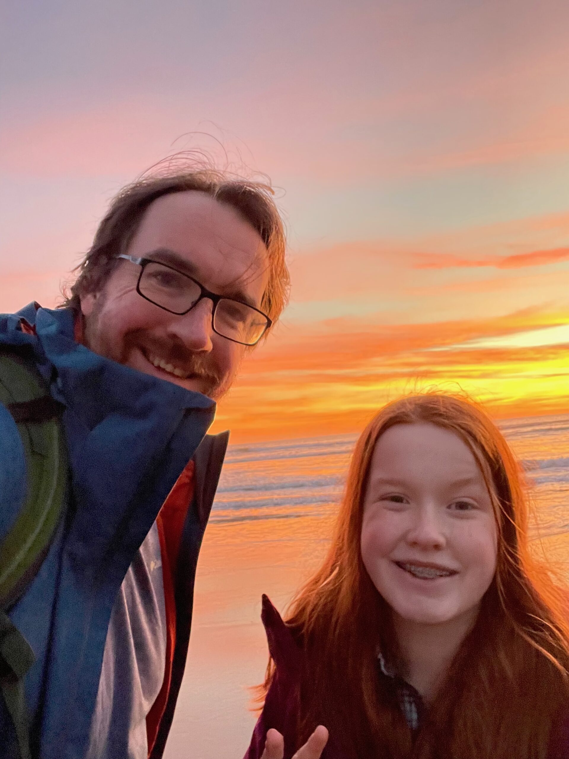 Cameron and myself pose for a photo at sunset at Cape Kiwanda. The sky and ocean are both bright red and orange from the sunset light and the color is washing over us. 